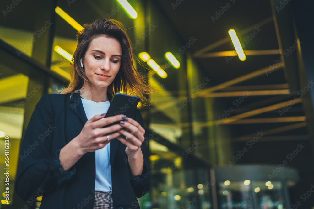 Successful female banker using smartphone outdoors while standing near his office background yellow neon lights, young woman professional manager working on mobile device near skyscraper at night - obrazy, fototapety, plakaty 