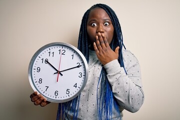 Poster - Young african american plus size woman with braids doing countdown holding big clock cover mouth with hand shocked with shame for mistake, expression of fear, scared in silence, secret concept