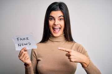 Wall Mural - Young beautiful brunette woman holding paper with fake news message over white background very happy pointing with hand and finger