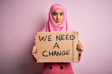 Wall Mural - Activist woman wearing pink muslim hijab holding banner with we need a change message with a confident expression on smart face thinking serious