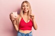 © Krakenimages.com - Young blonde woman holding piggy bank smiling with an idea or question pointing finger with happy face, number one
