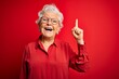 Senior beautiful grey-haired woman wearing casual shirt and glasses over red background pointing finger up with successful idea. Exited and happy. Number one.