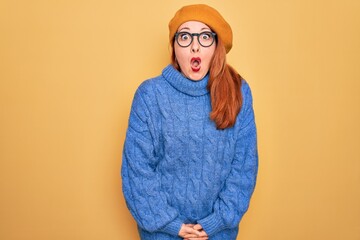 Wall Mural - Young beautiful redhead woman wearing french beret and glasses over yellow background afraid and shocked with surprise expression, fear and excited face.