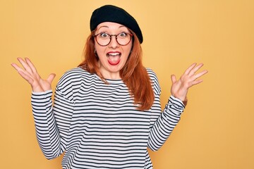 Wall Mural - Beautiful redhead woman wearing striped t-shirt and french beret over yellow background celebrating crazy and amazed for success with arms raised and open eyes screaming excited. Winner concept