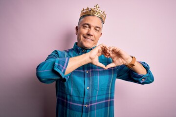 Poster - Middle age handsome grey-haired man wearing golden crown of king over pink background smiling in love showing heart symbol and shape with hands. Romantic concept.