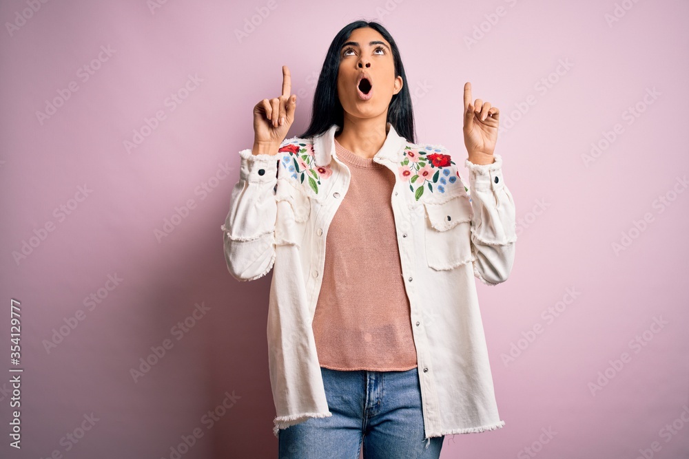 Young beautiful hispanic fashion woman wearing casual sweater over pink background amazed and surprised looking up and pointing with fingers and raised arms.