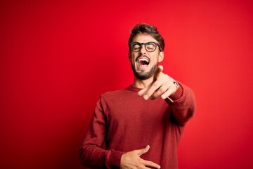 Wall Mural - Young handsome man with beard wearing glasses and sweater standing over red background laughing at you, pointing finger to the camera with hand over body, shame expression