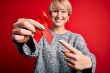 Poster - Young blonde woman with short hair holding hiv and aids awareness red ribbon very happy pointing with hand and finger