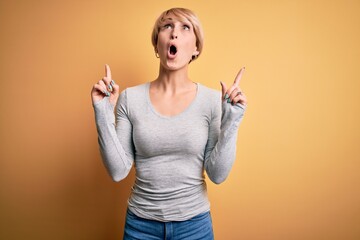 Wall Mural - Young blonde woman with modern short hair standing over yellow background amazed and surprised looking up and pointing with fingers and raised arms.