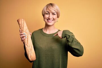 Poster - Young blonde woman with short hair holding fresh bread baguette over yellow background with surprise face pointing finger to himself