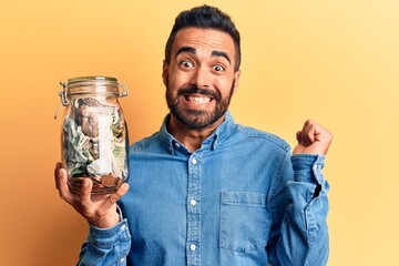 Young hispanic man holding jar with savings screaming proud, celebrating victory and success very excited with raised arm