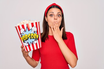 Young beautiful brunette woman eating pack of popcorns snack over isolated white background cover mouth with hand shocked with shame for mistake, expression of fear, scared in silence, secret concept