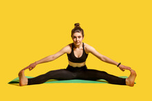 Gymnast Woman With Hair Bun In Tight Sportswear Sitting On Mat With Spread Legs, Touching Toes Stretching Muscles, Doing Sports Flexibility Exercises. Indoor Studio Shot Isolated On Yellow Background