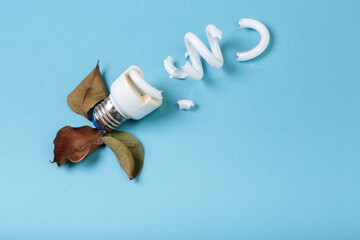 Broken fluorescent spiral tube lamp energy-saving lamp surrounded by dead ficus leaves on a blue background. Concept of ecology pollution