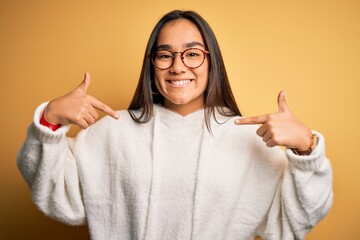 Young beautiful asian woman wearing casual sweater and glasses over yellow background looking confident with smile on face, pointing oneself with fingers proud and happy.