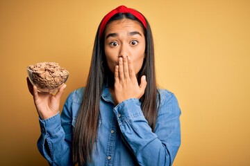 Sticker - Beautiful asian woman holding bowl with healthy corn flakes cereals over yellow background cover mouth with hand shocked with shame for mistake, expression of fear, scared in silence, secret concept