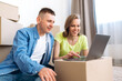 Handsome guy and attractive girl in a good mood shops online using a laptop. They moved to a new home. Happy couple in casual wear sitting on the floor among cardboard boxes