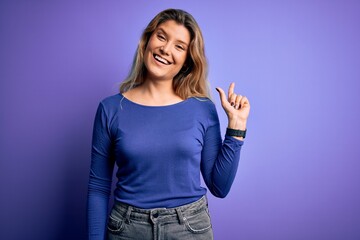 Poster - Young beautiful blonde woman wearing casual t-shirt over isolated purple background with a big smile on face, pointing with hand finger to the side looking at the camera.