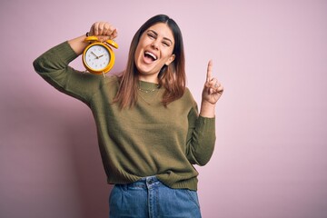 Poster - Young beautiful woman holding alarm clock standing over isolated pink background surprised with an idea or question pointing finger with happy face, number one