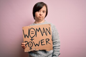 Sticker - Young down syndrome woman holding women power protest banner over pink background with a confident expression on smart face thinking serious