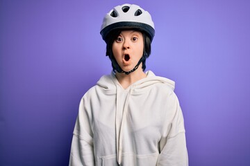 Wall Mural - Young down syndrome cyclist woman wearing security bike helmet over purple background afraid and shocked with surprise expression, fear and excited face.