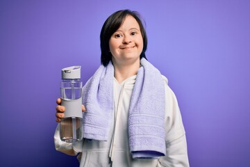 Canvas Print - Young down syndrome fitness woman training at gym holding water bottle and sport towel with a happy face standing and smiling with a confident smile showing teeth