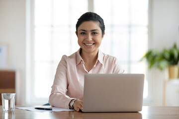 Head shot portrait young smiling indian woman sitting at table with computer. Happy hindu businesswoman professional looking at camera, spending time at workplace with laptop in modern office.