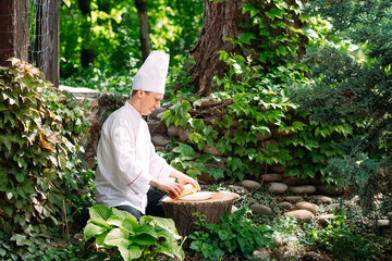 A restaurant chef in uniform rolls out dough on a stump in the woods.