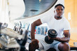 © twinsterphoto - Young African American man sitting and lifting a dumbbell close to the rack at gym. Male weight training person doing a biceps curl in fitness center