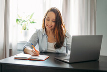 Adult Smiling Brunette Woman Doing Notes In Daily Book With Opened Laptop. She Study Something With Online Course At The Home