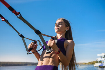 Pretty woman with sportwear doing crossfit push ups with trx fitness straps outdoors near the lake at daytime. Healthy lifestyle.