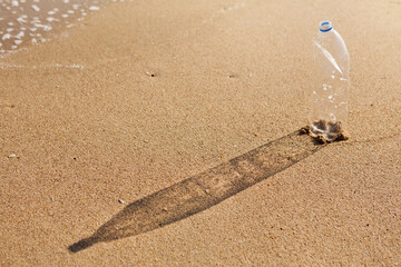 empty crumpled plastic bottle stands on the sea beach casting a long shadow, nature pollution concept