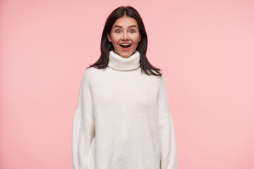 Wall Mural - Surprised young lovely brunette female with loose hair looking excitedly at camera with opened mouth and keeping hands along body while posing over pink background