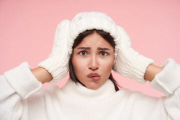 Wall Mural - Bewildered young green-eyed brunette female with loose hair keeping raised hands on her head while looking confusedly at camera, standing over pink background