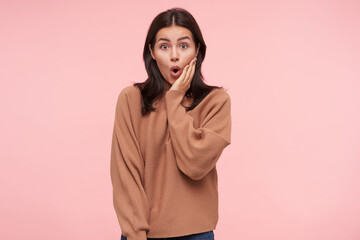 Wall Mural - Surprised young pretty brunette female with loose hair holding raised palm on her cheek while looking amazedly at camera with wide eyes opened, isolated over pink background