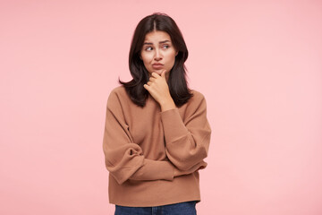 Wall Mural - Bewildered young attractive brown haired lady dressed in knitted roll-neck sweater touching her chin with raised hand and looking thoughtfully aside while posing over pink background