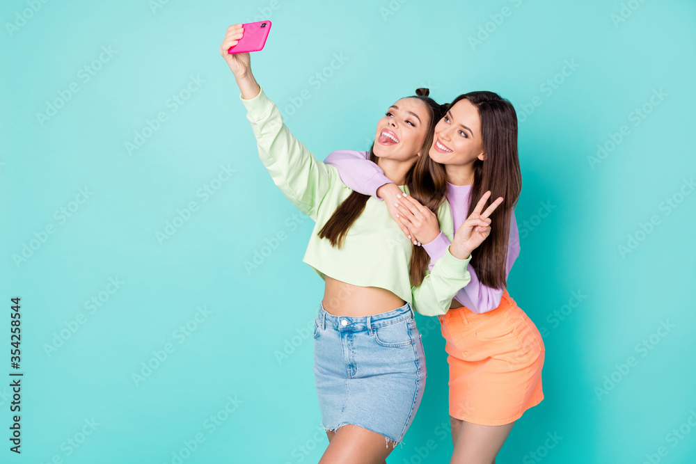 Photo of two pretty cheerful ladies best friends hold telephone take selfies showing v-sign symbol wear cropped sweaters naked belly short skirts isolated pastel teal color background - obrazy, fototapety, plakaty 