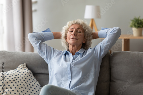 Old 70s woman dreamer seated on couch relaxing meditating thinking positive thoughts putting hands behind head closed eyes breathing fresh air relish retired life free time at home, well-being concept