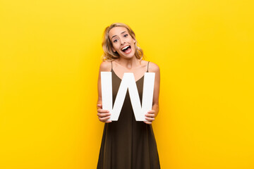 Wall Mural - young blonde woman surprised, shocked, amazed, holding the letter W of the alphabet to form a word or a sentence.