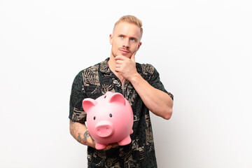 Wall Mural - young blonde man thinking, feeling doubtful and confused, with different options, wondering which decision to make with a piggy bank