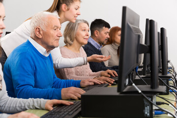 Young female teacher helping mature people to use computer