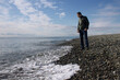 © Ekaterina - A man dressed in blue jeans and a jacket stands on a rocky beach and looks at the edge of the surf, thinking about something.