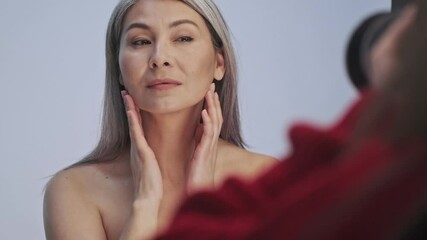 Wall Mural - A beautiful elderly half-naked woman is posing to photographer isolated over gray background in studio