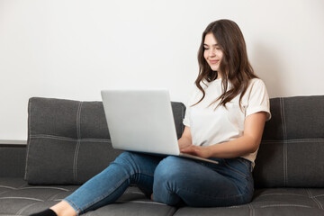 young beautiful brunette woman texting message in her laptop sitting on the sofa in her appartment, feeling cozy at home