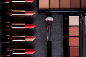 Lipsticks lie on a dark surface, next to which are palettes with shadows and a brush. Cosmetic theme.