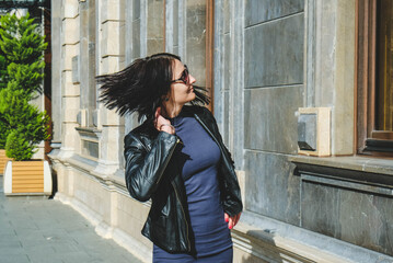 Wall Mural - Brunette woman with a positive smile in a dress and leather black jacket shake hair in street and enjoys a sunny spring day. Girl travel in Batumi, Georgia. Old city at background.