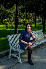 Wall Mural - Nice, brunette woman in a dress sits on white bench in the street. Enjoys a sunny spring day. Girl traveling in Batumi, Georgia. Green park at background.