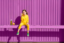 Little Girl Dressed In Yellow Sitting On Bar In Front Of Purple Background Playing With Banana