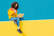 © VISTA by Westend61 - Happy young woman with coffee to go sitting on yellow wall using laptop