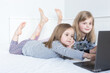 Two white child girls watching cartoons in a laptop while lying on the bed. Gray clothes, bare feet. Bright interior. Blurred background. Selective focus.